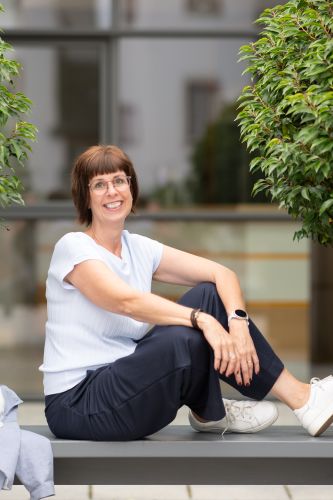 Monika Biewer sitzt auf einer Bank, Beine sind auch auf der Bank und sie lächelt