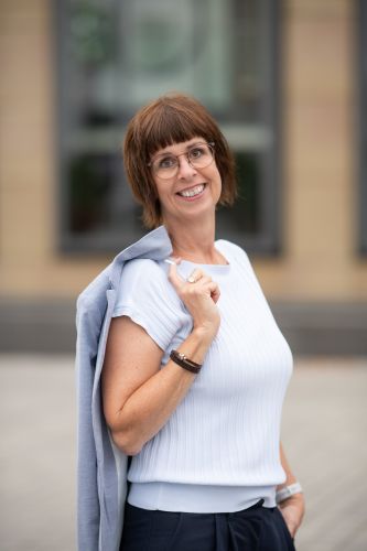 Monika Biewer steht aufrecht, Blazer locker über die Schulter festhaltend, die andere Hand in der Hosentasche